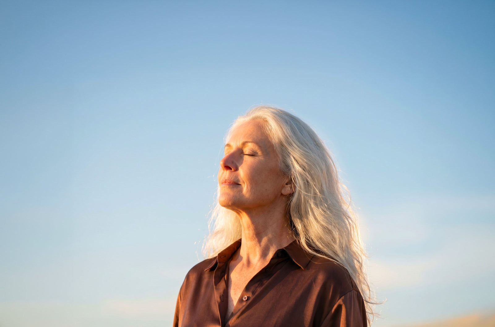 Woman with long silver hair standing outdoors with eyes closed, calm against a blue sky
